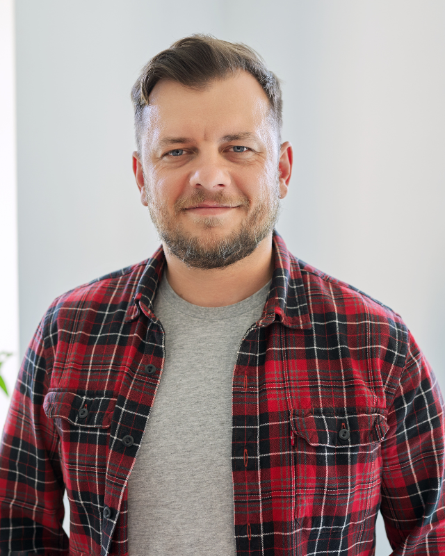 A confident man with a beard smiling gently at the camera, wearing a grey t-shirt under an open red and black checkered shirt, with a bright, airy background.